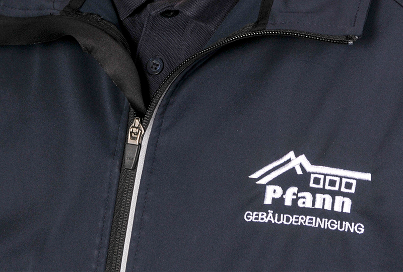 Nahaufnahme einer dunklen Jacke mit sichtbarem Reißverschluss und einem aufgestickten Logo der Pfann Gebäudereinigung mit einem stilisierten Dach und Fenstern über dem Text.